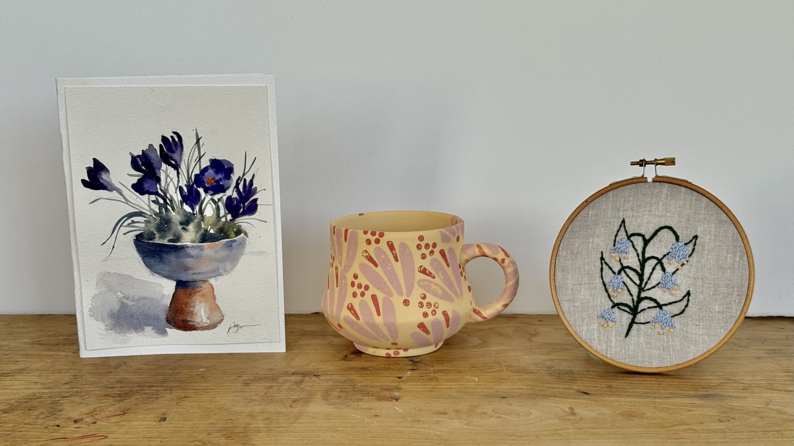 Projects to choose from a a maker's circle art bar paint and sip: watercolor painted greeting card of purple crocuses, underglaze tatoo decorated ceramic mug with pink botanical pattern, and embroidery hoop with blue wildflower design displayed on wood table