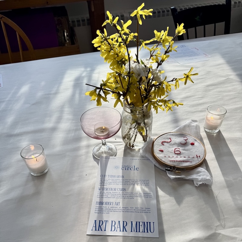 Wine and Art Night, New Jersey. Art Bar table setting at A Maker's Circle in Pottersville NJ with forsythia flower arrangement, candles, coupe glass, embroidery hoop, and Art Bar menu