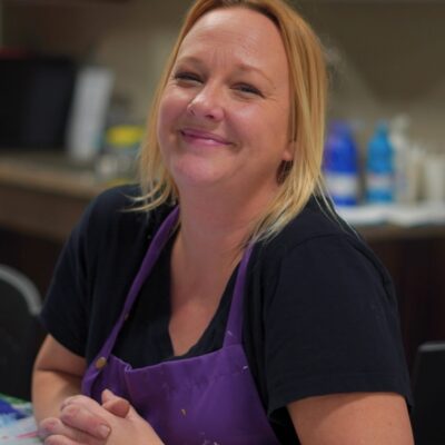 Vanessa Lopez, kids art instructor at A Maker's Circle in Pottersville NJ, smiling in a paint-splattered purple apron in the art studio