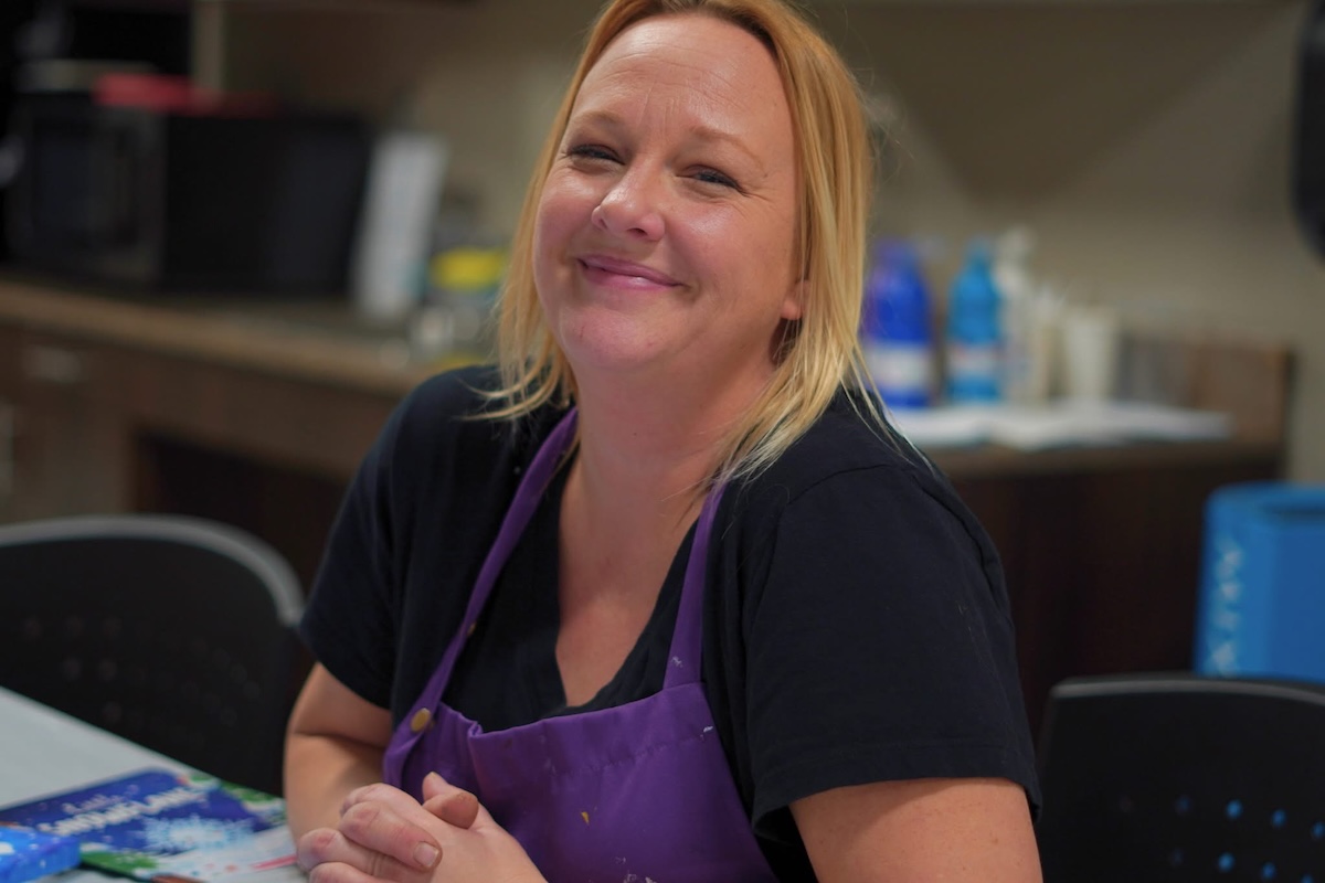 Vanessa Lopez, kids art instructor at A Maker's Circle in Pottersville NJ, smiling in a paint-splattered purple apron in the art studio