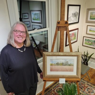 Jane Ramsey, watercolor and drawing instructor at A Maker's Circle in Pottersville NJ, standing next to her framed watercolor landscape paintings in her gallery
