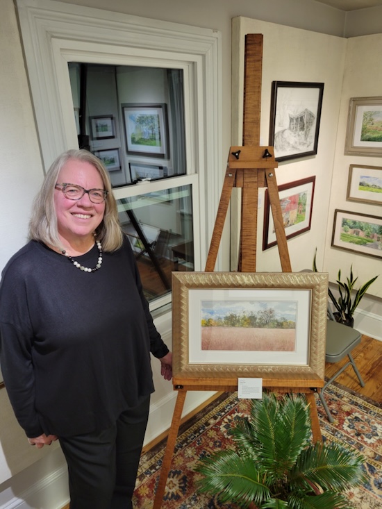 Jane Ramsey, watercolor and drawing instructor at A Maker's Circle in Pottersville NJ, standing next to her framed watercolor landscape paintings in her gallery