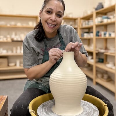 Lisette Bedoya, pottery instructor at A Maker's Circle in Pottersville NJ, smiling at the pottery wheel while throwing a large clay vase