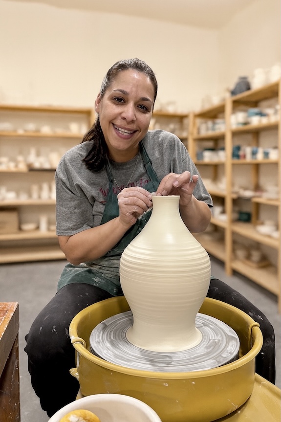Lisette Bedoya, pottery instructor at A Maker's Circle in Pottersville NJ, smiling at the pottery wheel while throwing a large clay vase